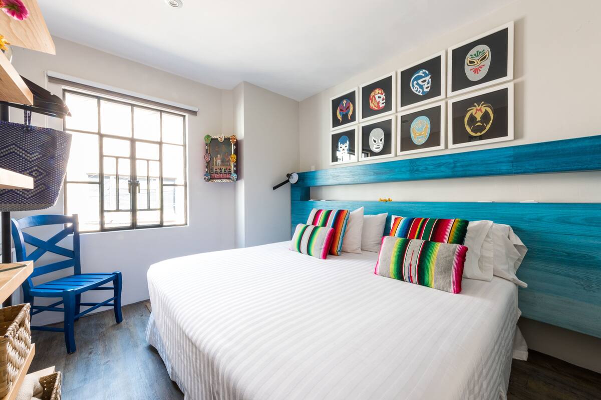 Un dormitorio con una cama grande, almohadas coloridas, un cabecero azul y máscaras enmarcadas en la pared.