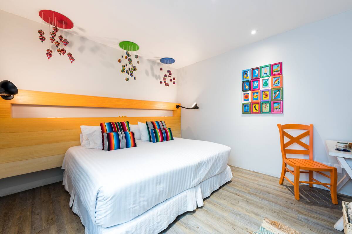 Un dormitorio con cabecera de madera, cama con ropa de cama a rayas, un cuadro de colores en la pared y suelo de madera.