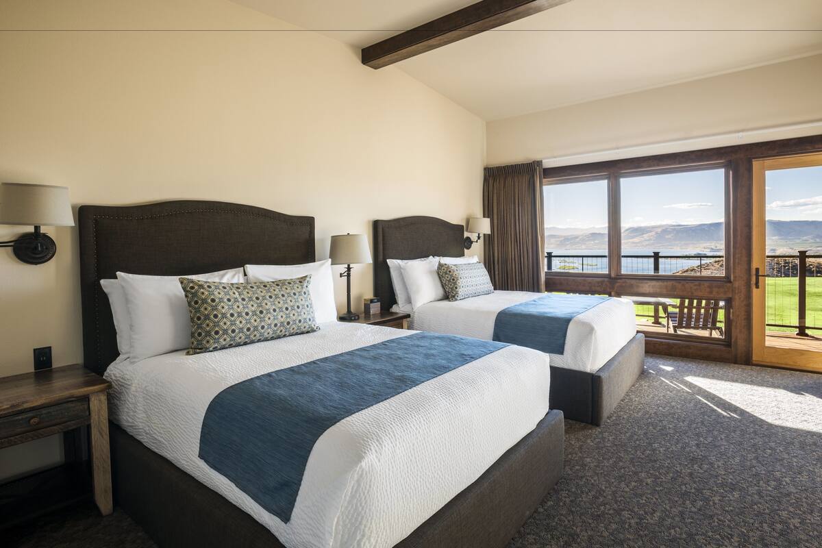 A hotel room with two beds, a large window with a view of mountains, and a balcony.