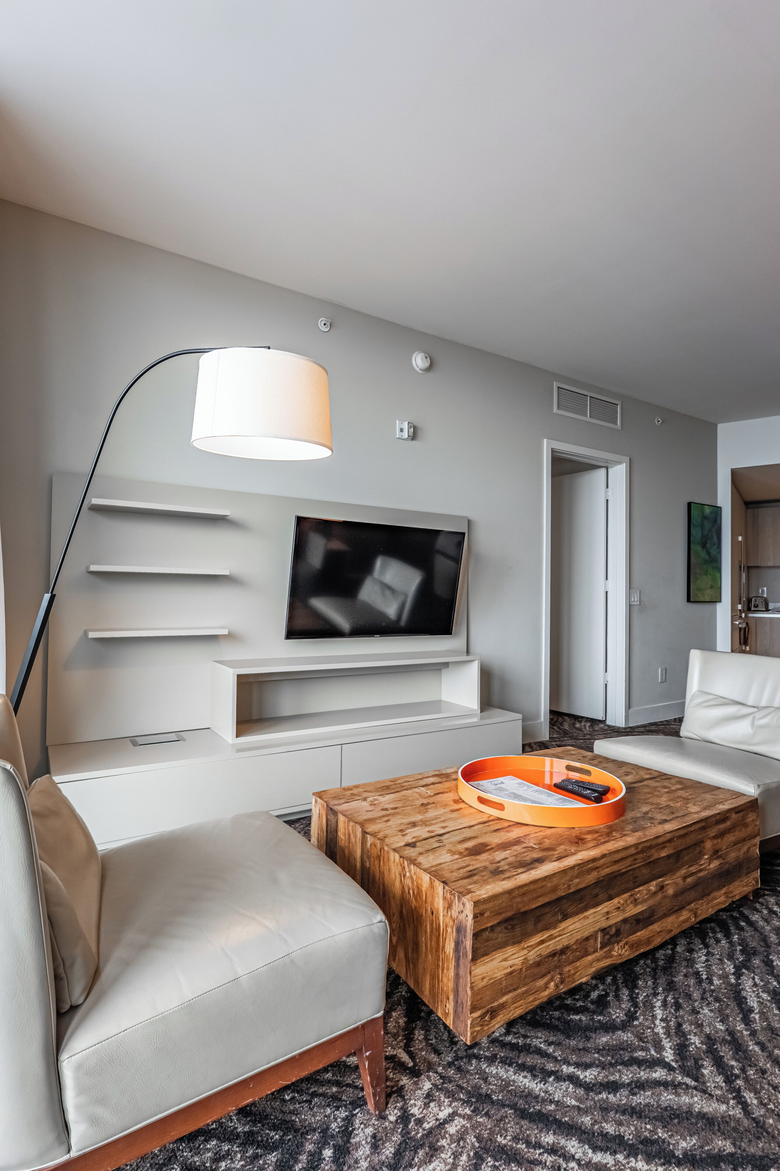A modern hotel room with a flat-screen TV mounted on the wall, a white sofa, a wooden coffee table with a tray, and a large floor lamp.