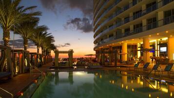 Zona de piscina hotelera con sillones, palmeras y vista al atardecer.