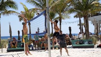Jogo de voleibol de praia com palmeiras e pessoas ao fundo.