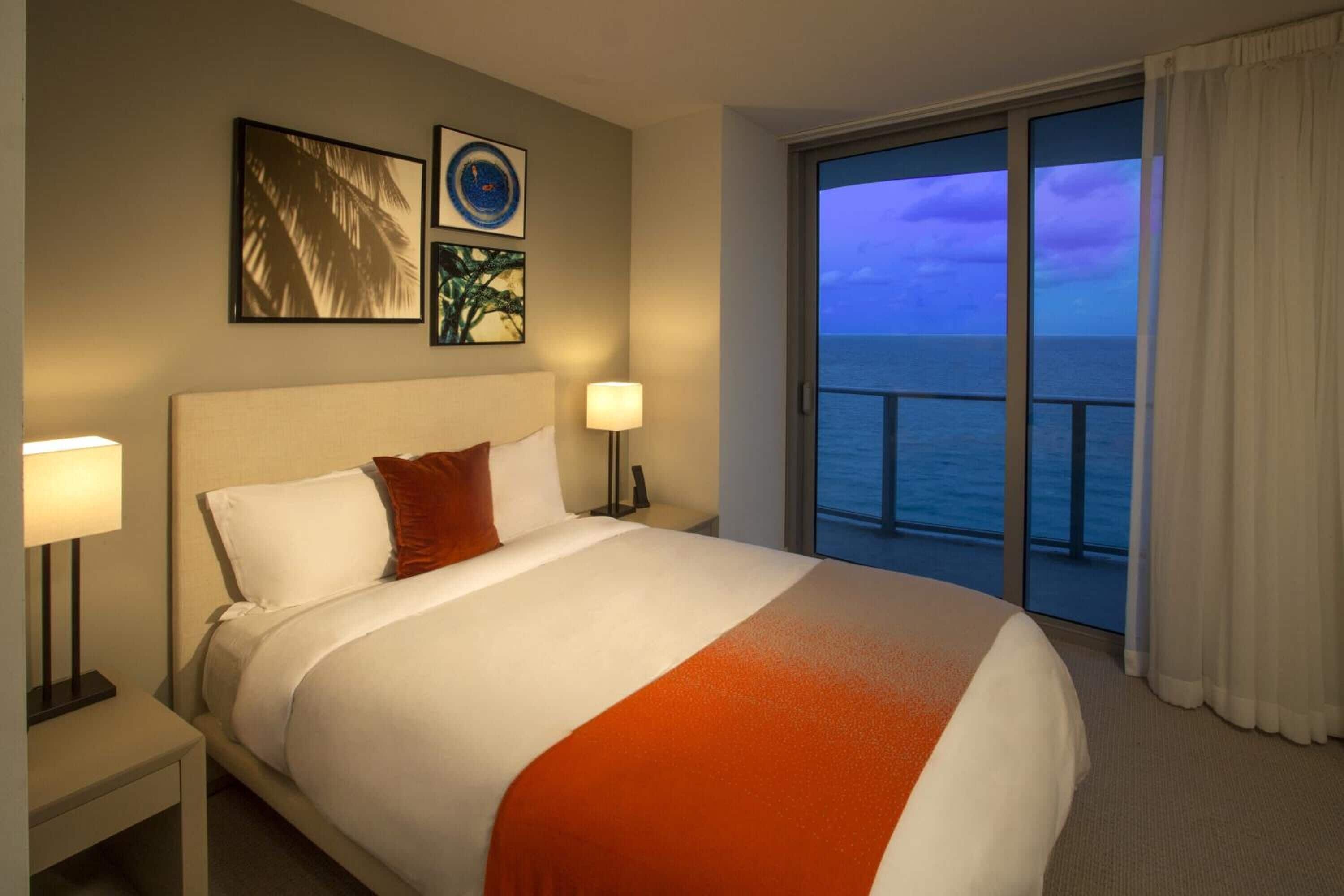 A hotel room with a large bed, two bedside lamps, a nightstand, and a view of the ocean through a balcony with curtains.