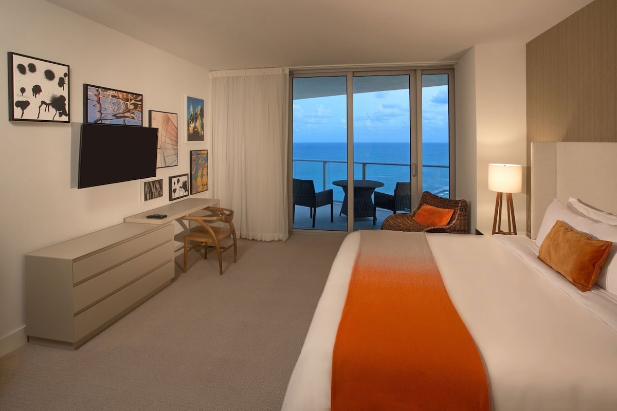 A hotel room with a bed, a desk, a chair, a TV, and a balcony with a view of the sea.