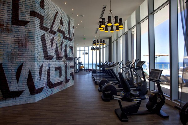 A gym with exercise equipment, large windows, and a brick wall with graffiti. - Lyfe Resort & Residences (Hollywood)