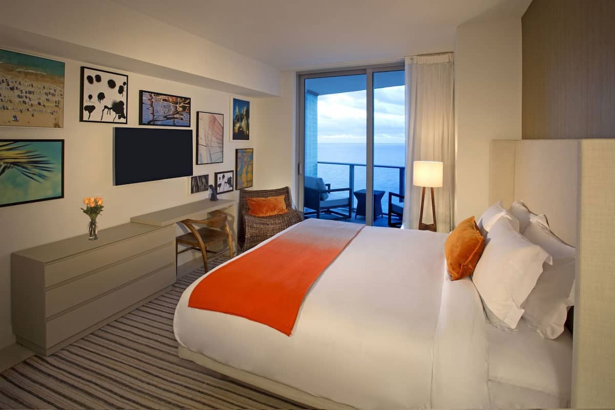 A hotel room with a bed, a desk, a chair, a balcony with a view of the sea, and artwork on the walls.