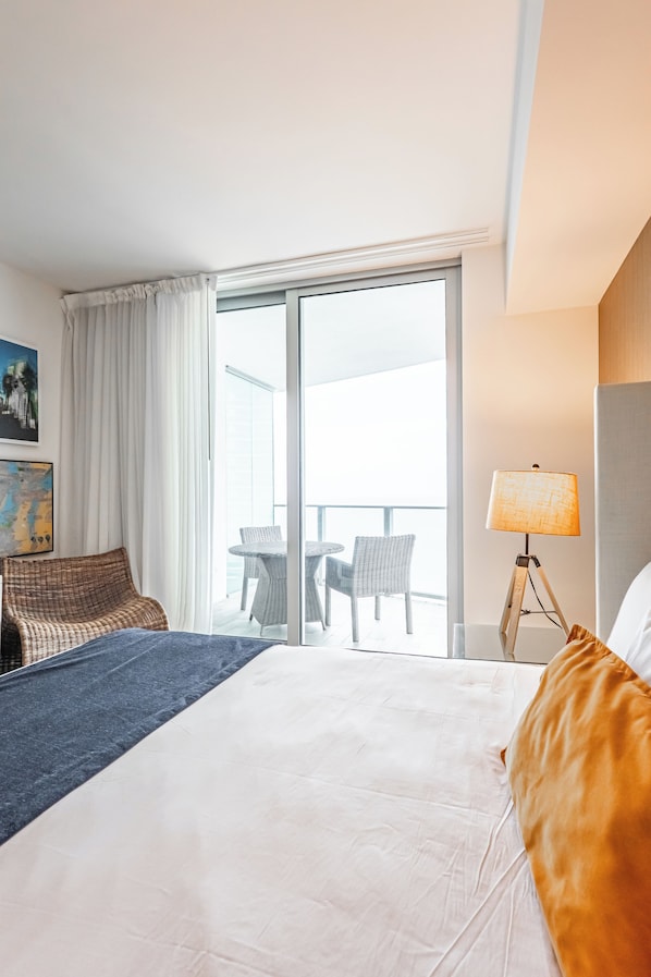 A hotel room with a bed, a chair, a table, a lamp, and a view of the outside. - Lyfe Resort & Residences (Hollywood)