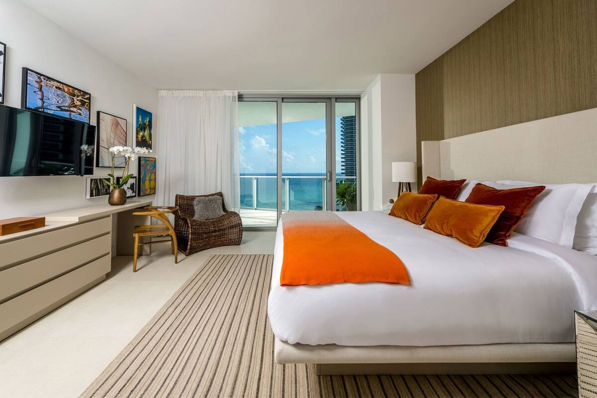 A hotel room with a large bed, a desk, a chair, a TV, and a balcony with a view of the ocean.