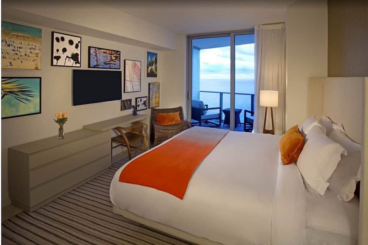 A hotel room with a bed, a desk, a chair, a lamp, and a view of the sea.