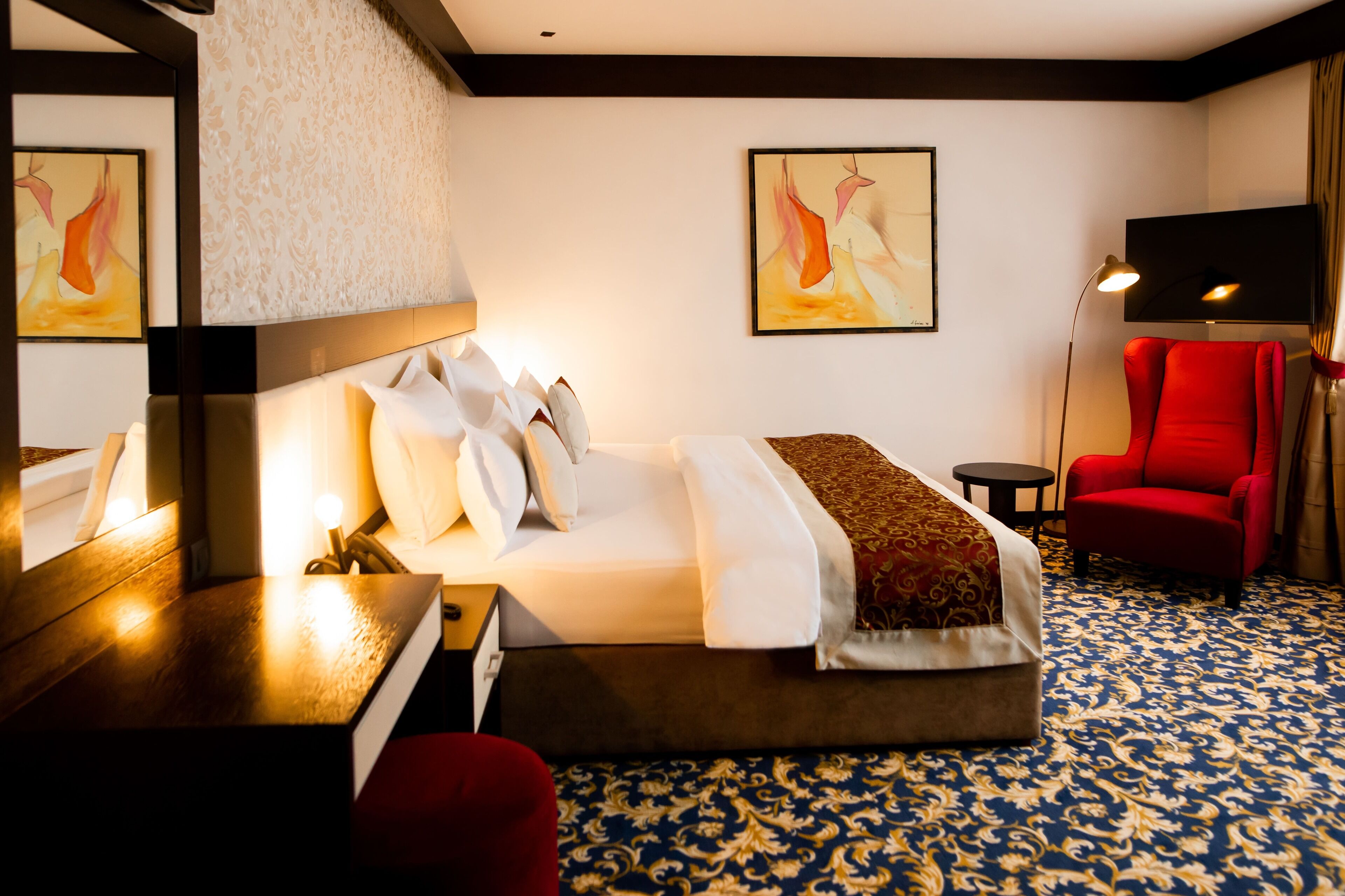 A hotel room with a bed, a red armchair, a small table, and a mirror.