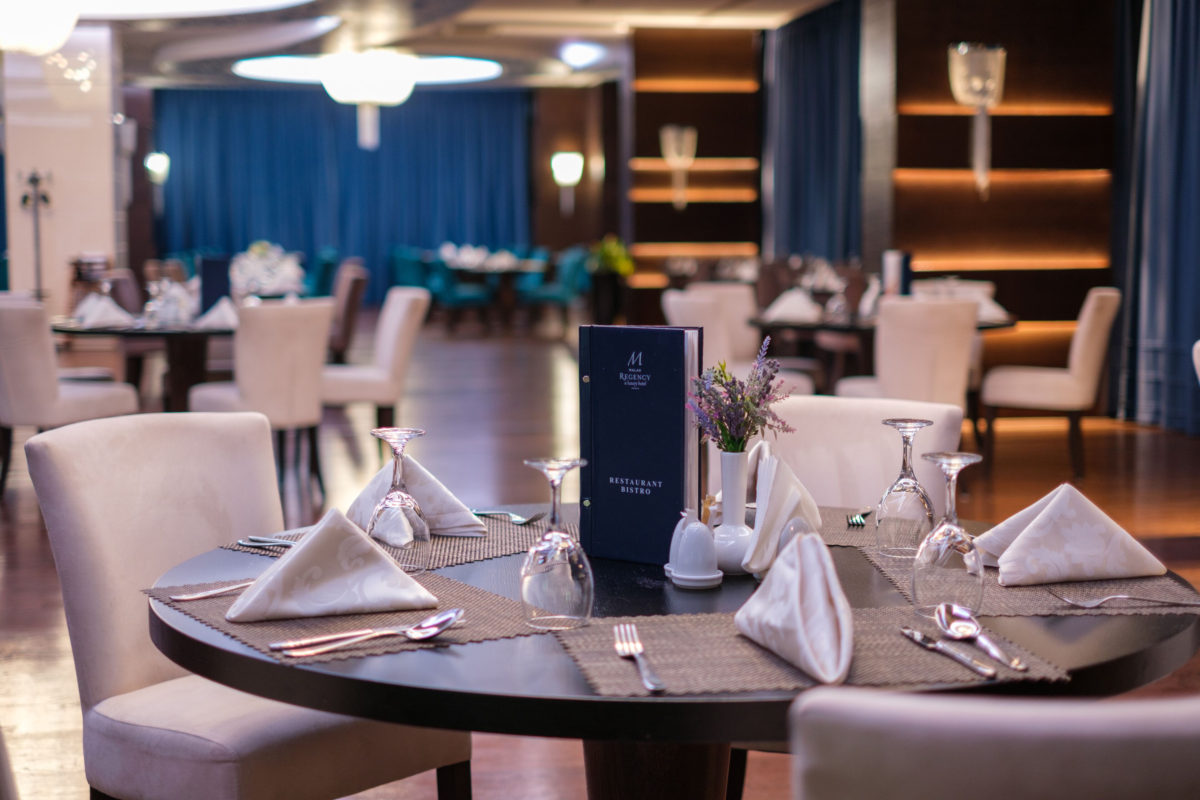 Un comedor con mesas redondas dispuestas para una comida, incluyendo vajilla, cubiertos y cristalería.