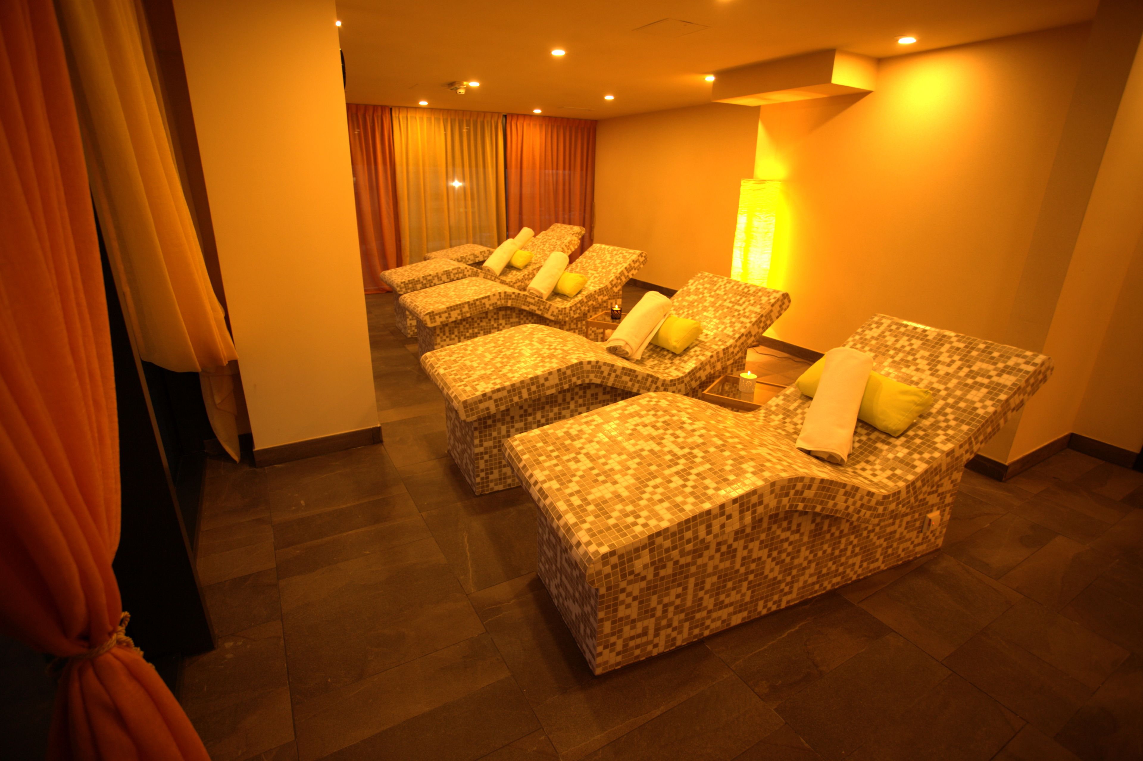 A room with four massage chairs, each with a towel and pillow. The room has a tiled floor and a warm, yellow-lit ambiance.