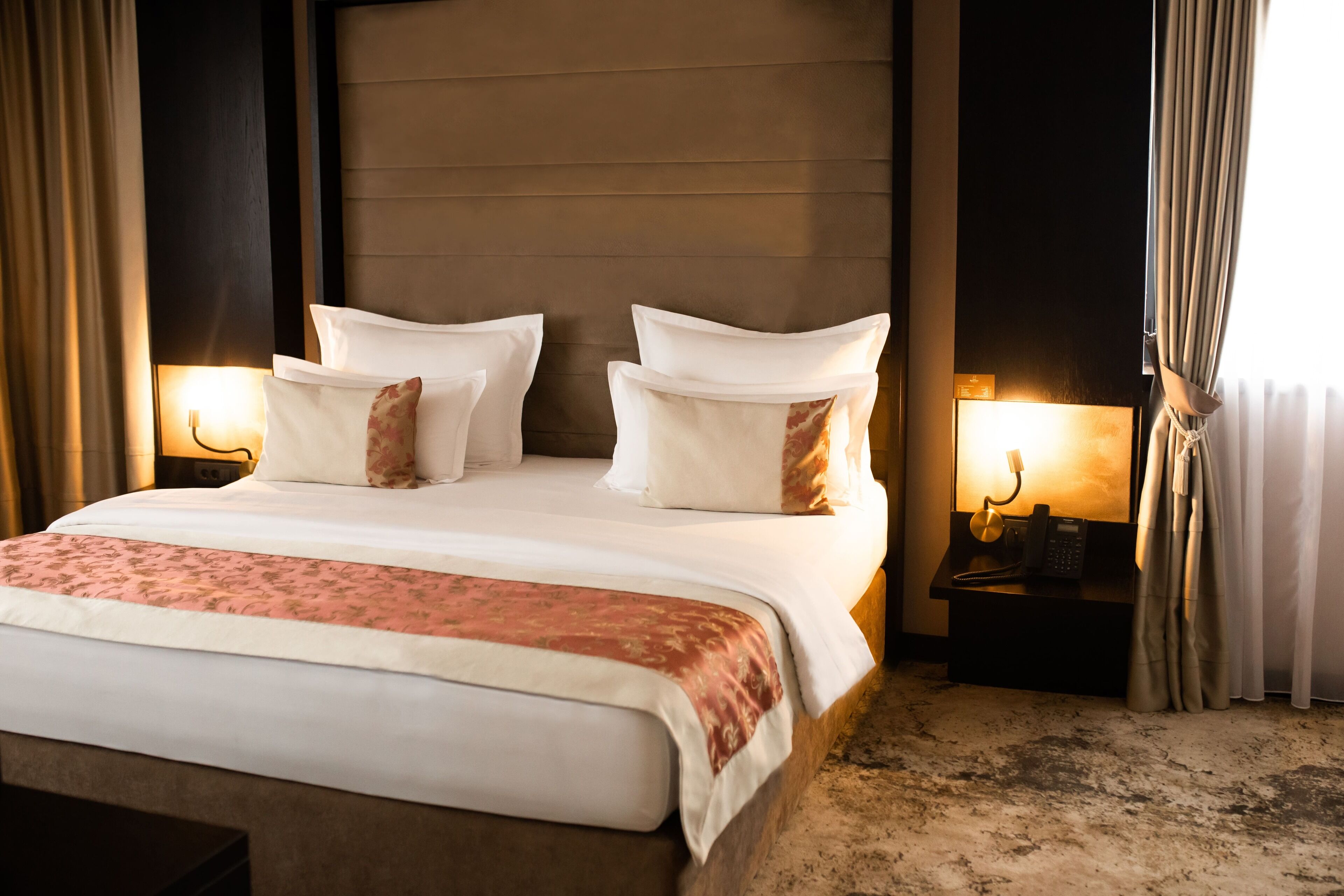 A hotel room with a large bed, two bedside tables with lamps, and a view of the outside through curtains.