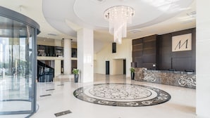 A spacious lobby with a circular patterned floor, a grand chandelier, and a reception desk with a large 'M' sign.