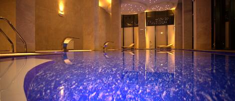 A large indoor swimming pool with a blue marble-like floor, a seating area with chairs, and a ceiling with small lights.