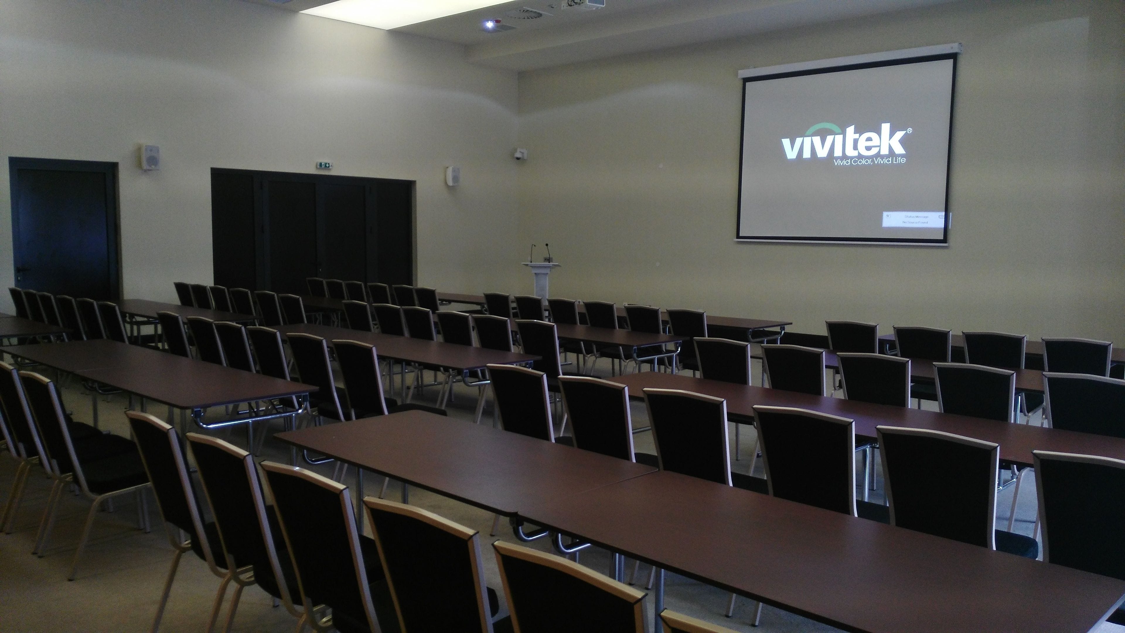 A conference room with rows of tables and chairs, a projector screen with 'vivitek' branding, and a podium.