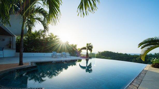 An infinity pool with a wooden deck and palm trees, surrounded by a modern building and lush greenery. - Casa Phil (Las Terrenas)