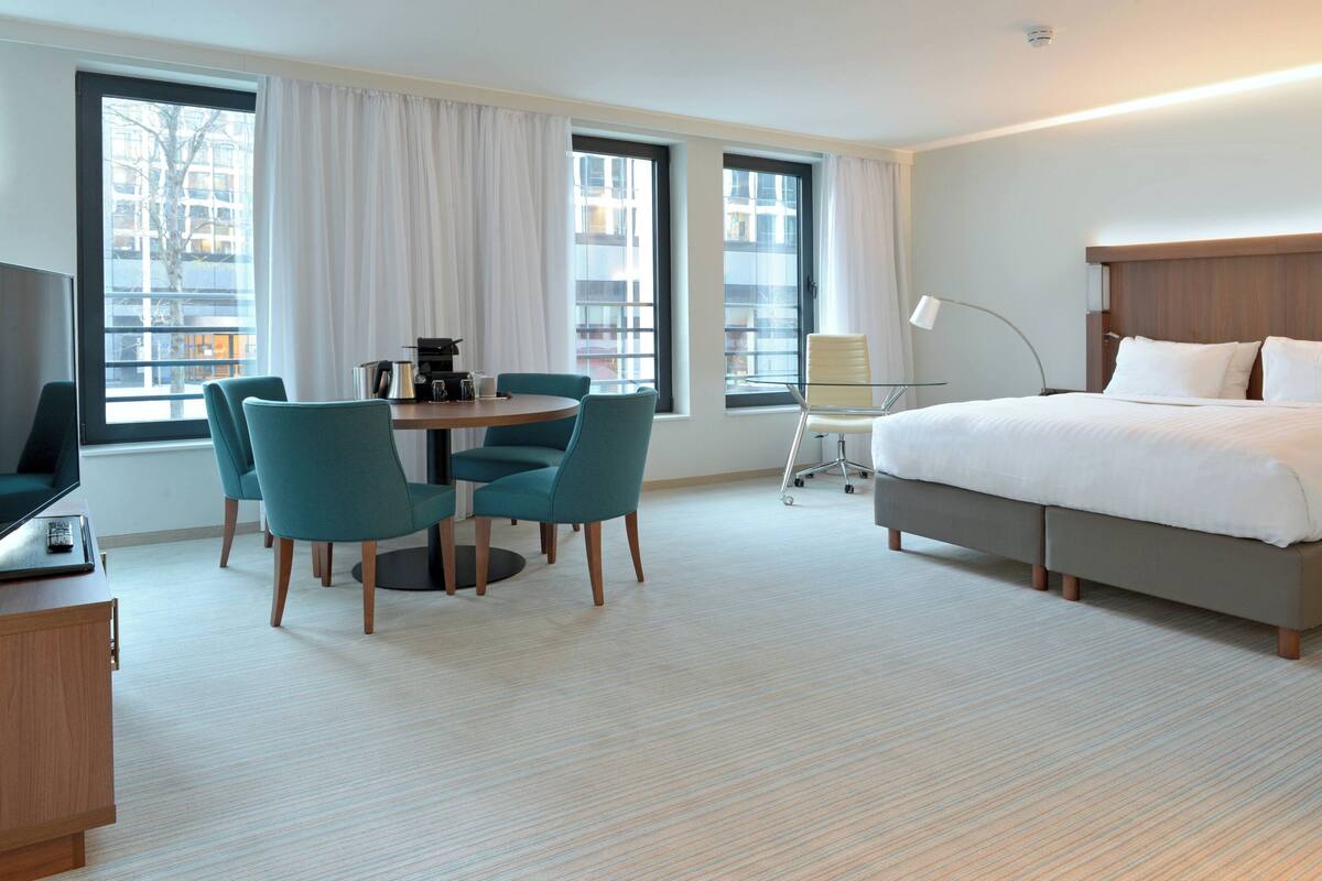 A hotel room with a bed, a desk with a chair, a small table with chairs, and large windows.