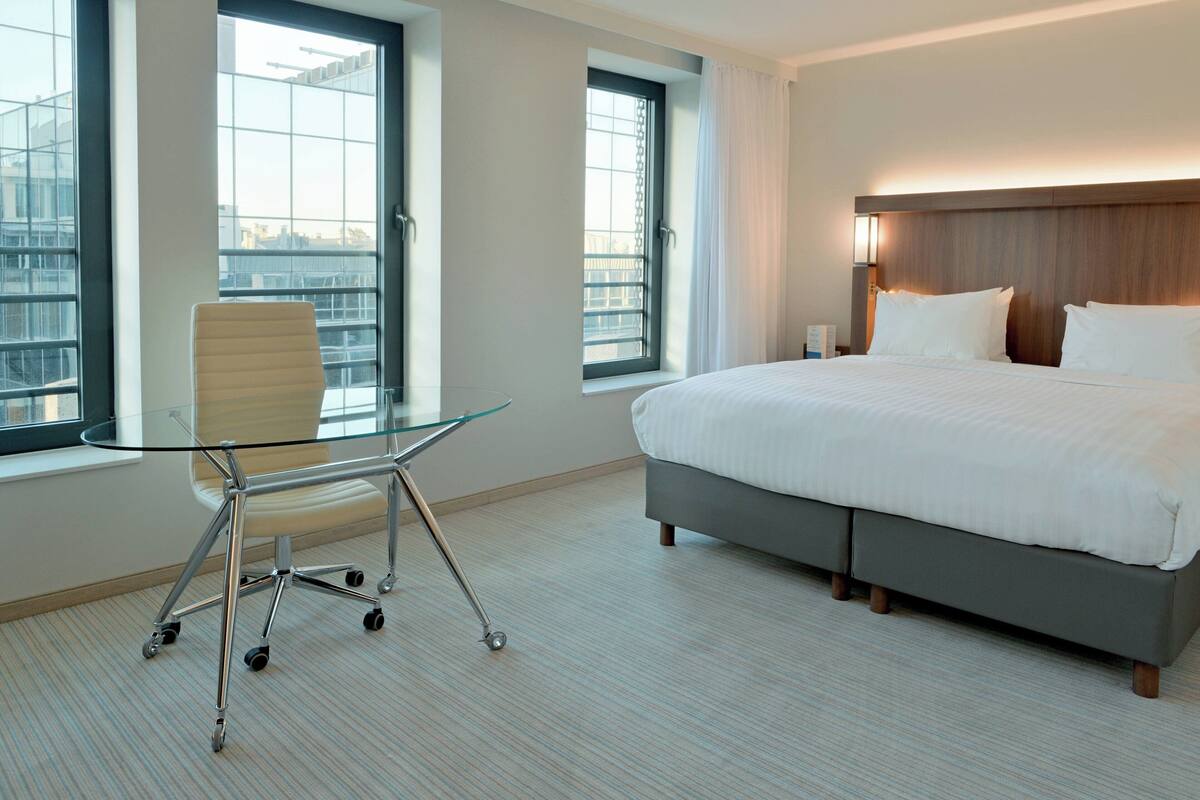 A modern hotel room with a large bed, a glass table with a chair, and large windows offering city views.