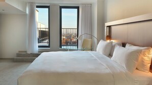 Habitación de hotel con una cama grande, una mesita de vidrio, una silla y vistas a la ciudad desde un balcón.