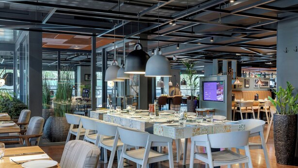 Un restaurante moderno con una larga mesa compartida, sillas blancas, iluminación colgante y una pantalla de televisión visible.
