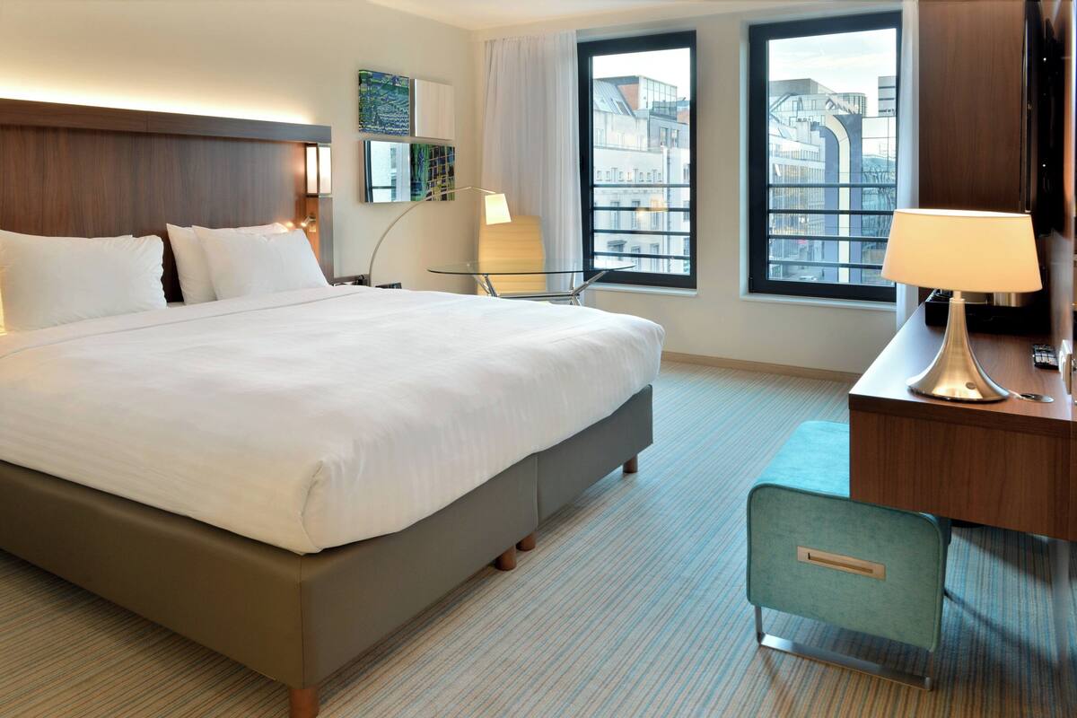 A hotel room with a large bed, a desk with a lamp, a chair, and a view of buildings through the window.