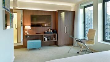 A hotel room with a bed, a desk with a chair, a TV, a minibar, and a large window.