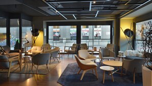 Un área de descanso moderna con diversas opciones de asientos, incluyendo sofás y sillones, dispuestos alrededor de una mesa de centro. Amplias ventanas ofrecen vista a la calle.
