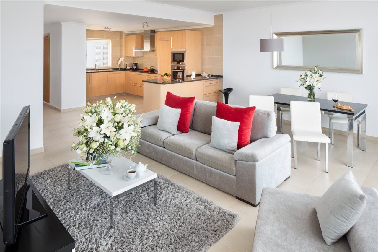A modern living room with a grey sofa, red and white cushions, a coffee table, and a TV. There is a kitchen in the background with wooden cabinets and a dining area with a table and chairs.