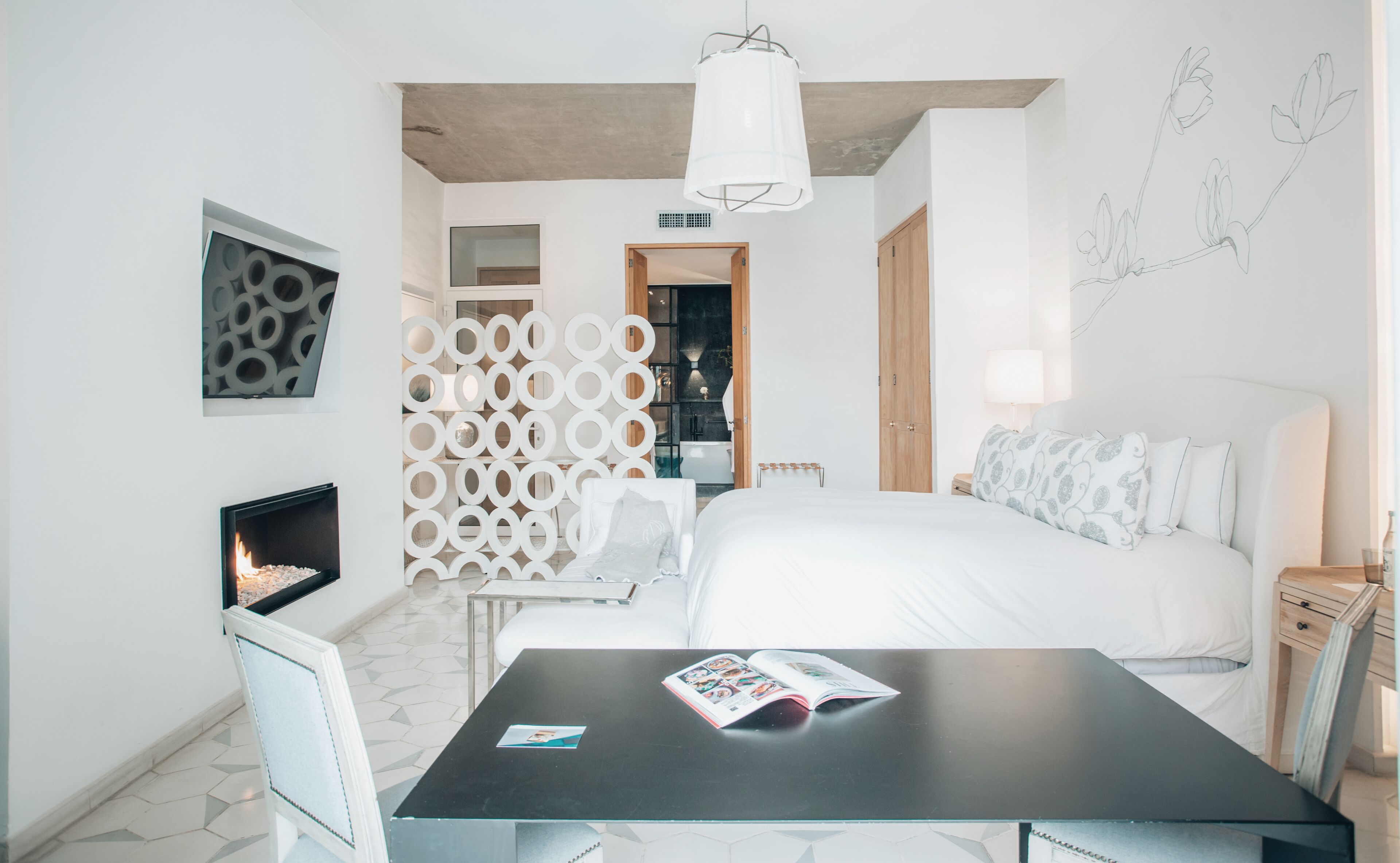 A modern hotel room with a bed, a desk, a chair, a fireplace, and a bathroom visible through a glass partition.
