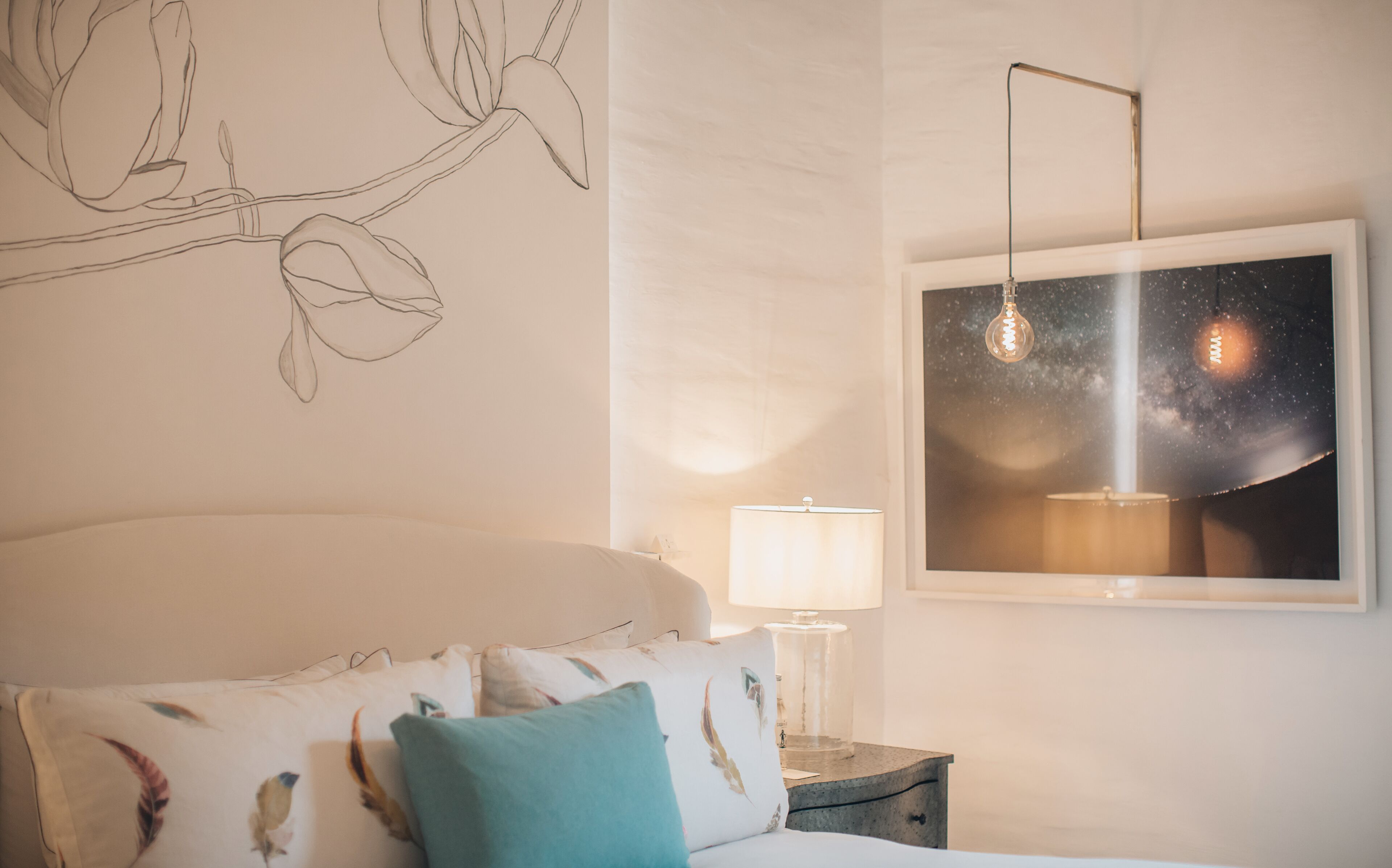 A bedroom with a bed, pillows, a nightstand, a lamp, a wall art of a night sky, and a branch with leaves.