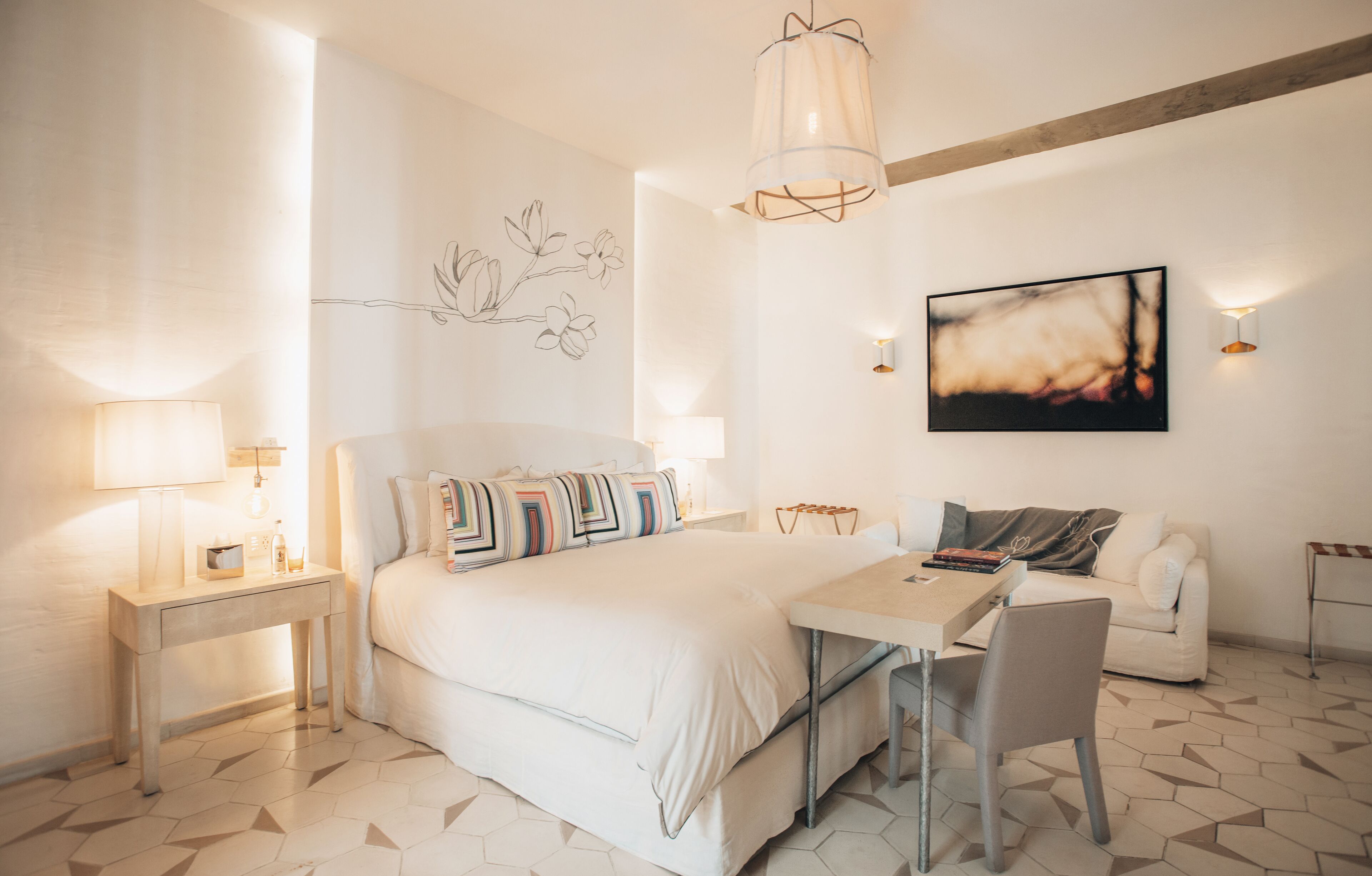 A modern bedroom with a large bed, bedside tables, a desk with a chair, a hanging light fixture, and a wall-mounted abstract painting.