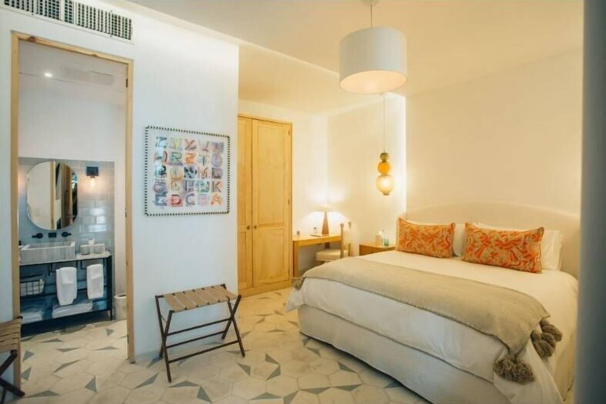 Habitación de hotel con cama, mesita de noche, lámpara, espejo, armario y baño con lavabo y toallero.
