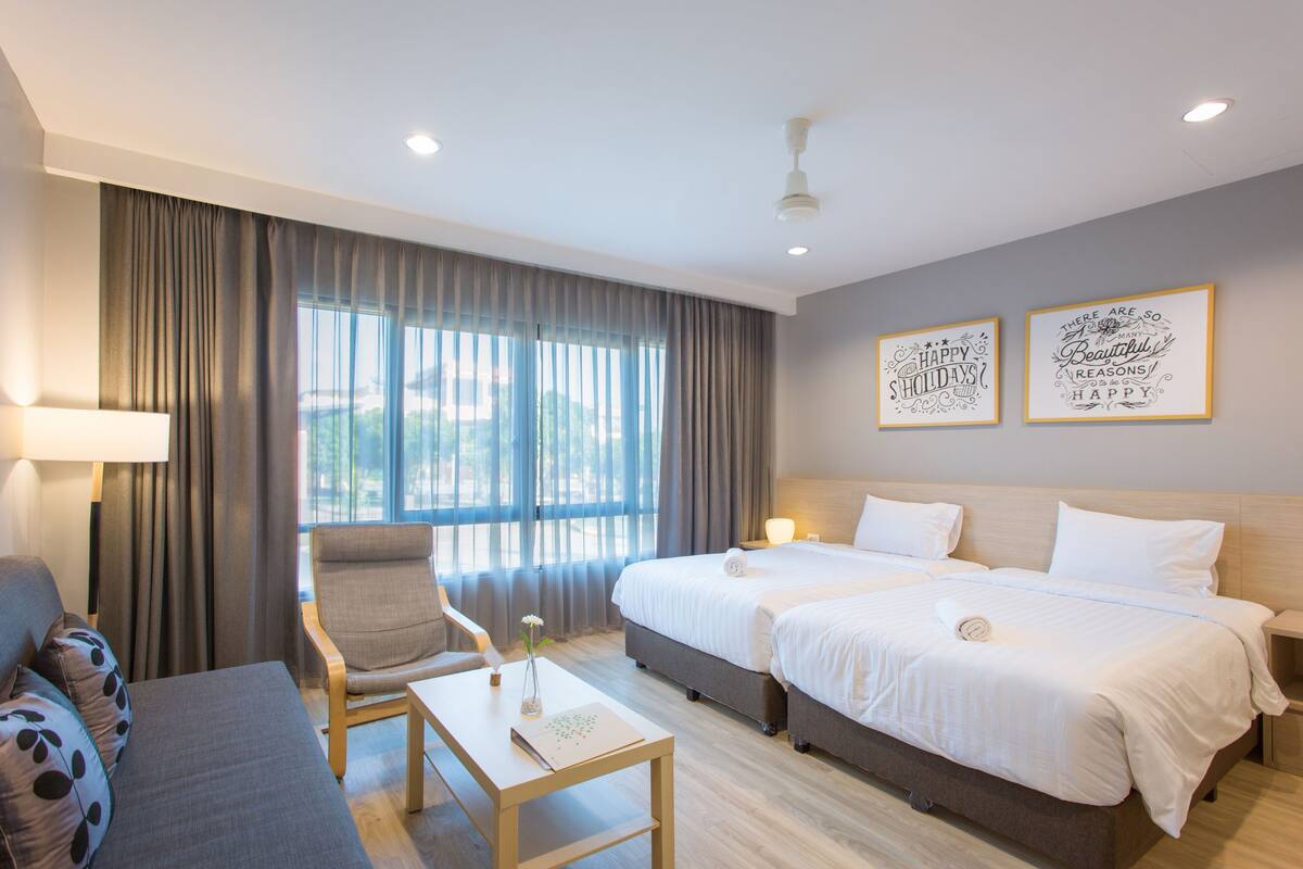 Quarto de hotel com duas camas, um sofá, uma mesa e uma cadeira. Há duas obras de arte nas paredes e uma janela com cortinas.