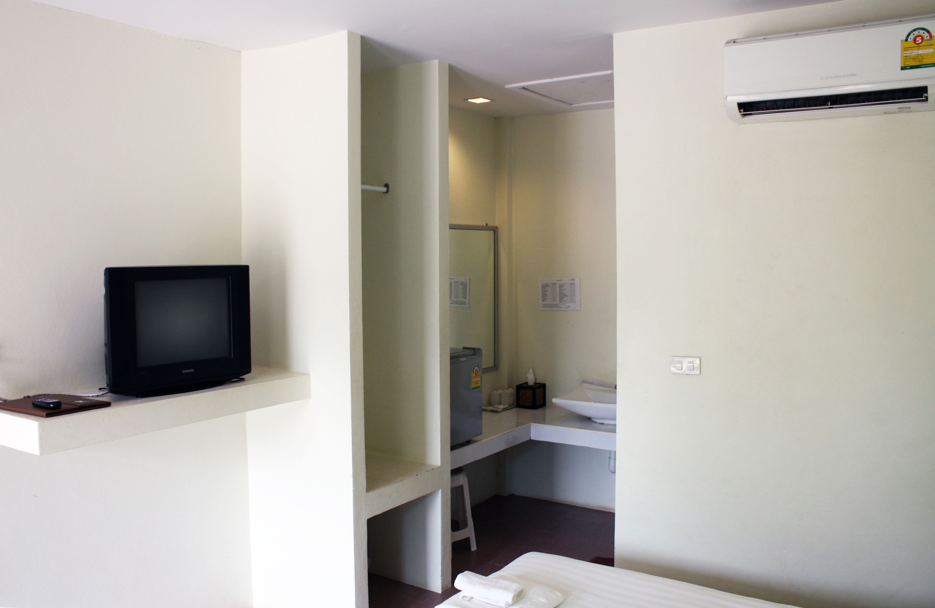 A hotel room with a television on a shelf, a small stool, a sink, and an air conditioning unit.