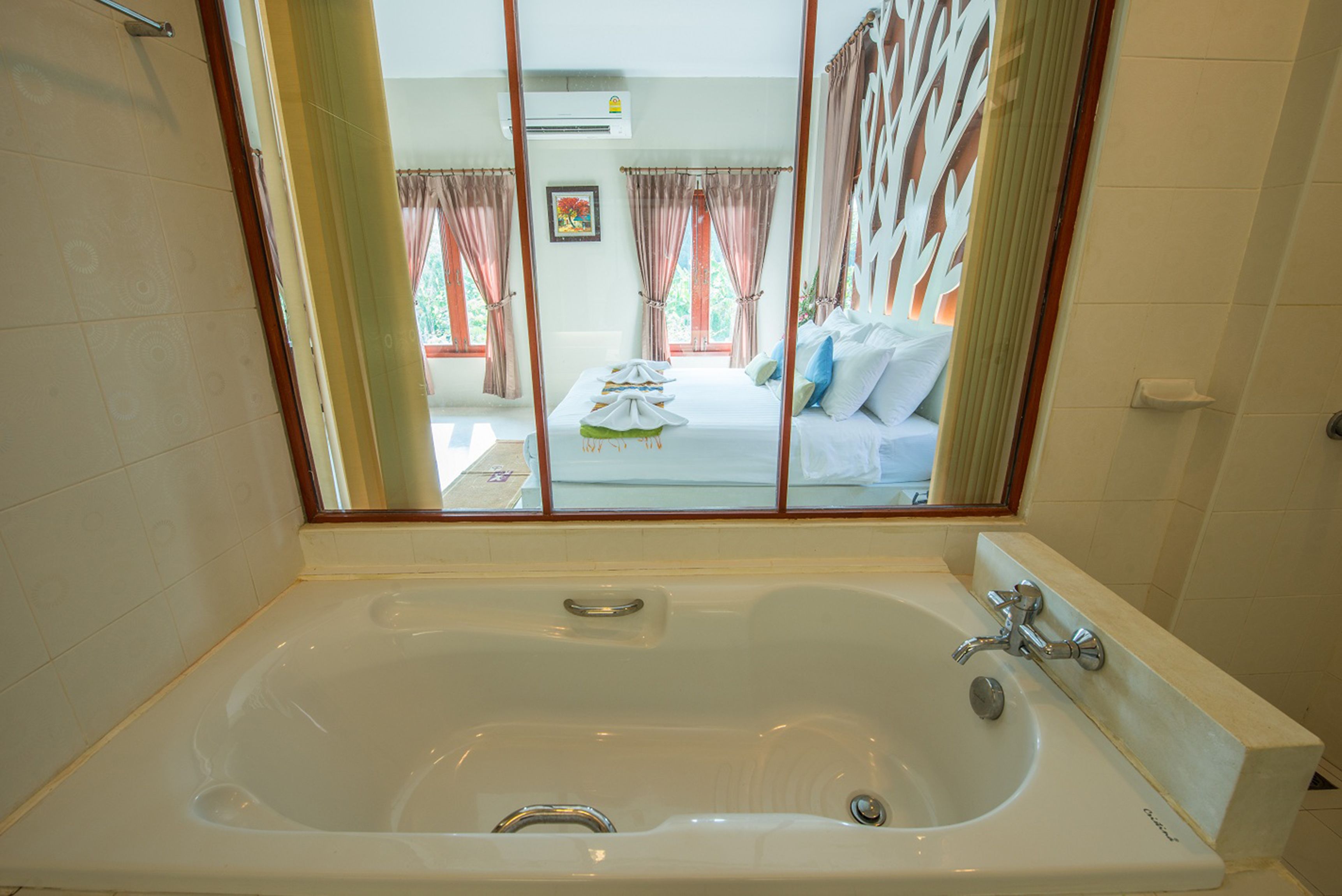 Une grande baignoire blanche, dotée d’une pomme de douche et d’un robinet, une fenêtre avec des rideaux, et un lit avec des oreillers et une couverture, visibles à travers la fenêtre.