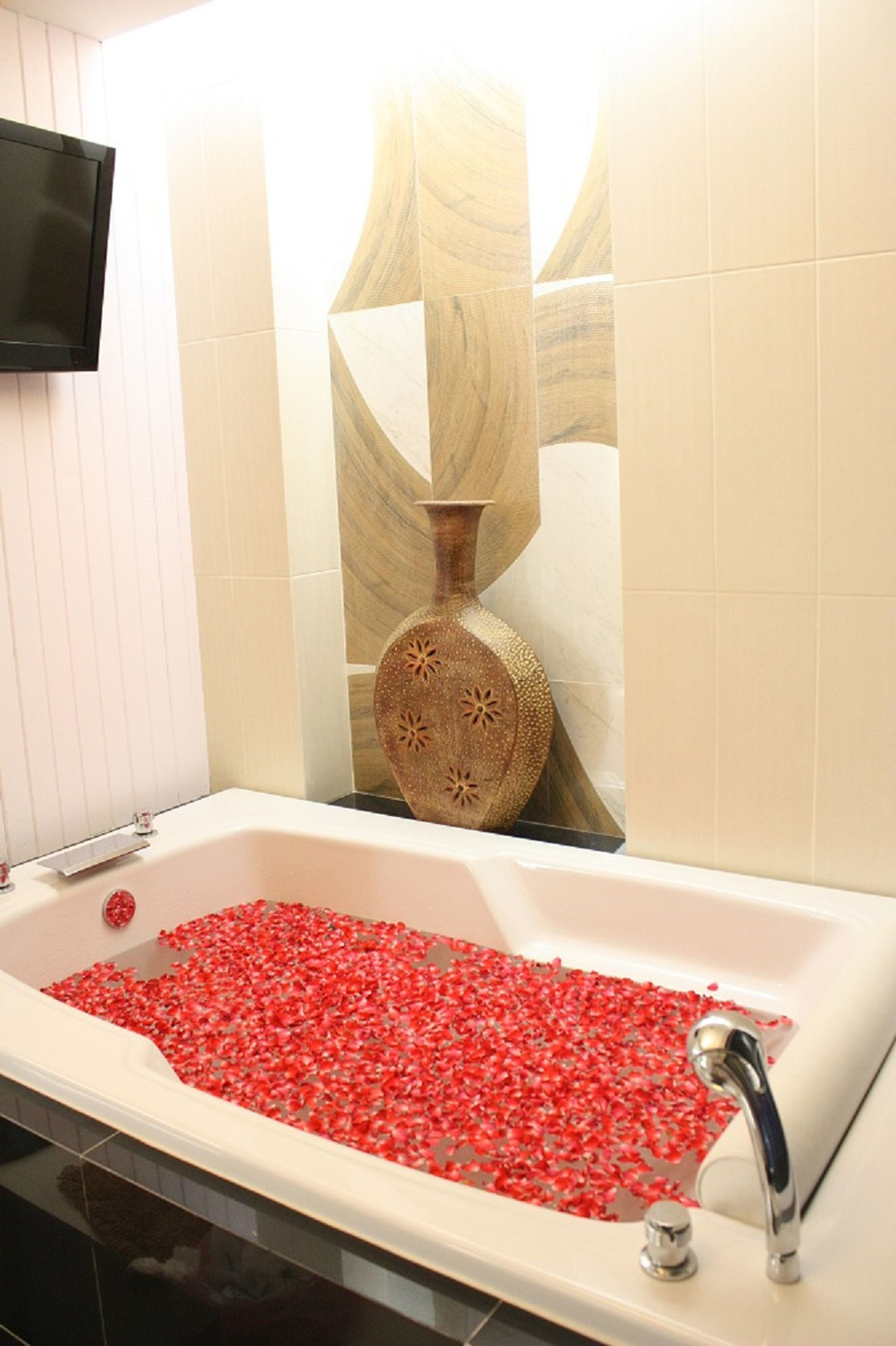 Una bañera llena de pétalos de rosa roja, un florero de madera y un televisor de pantalla plana montado en la pared.