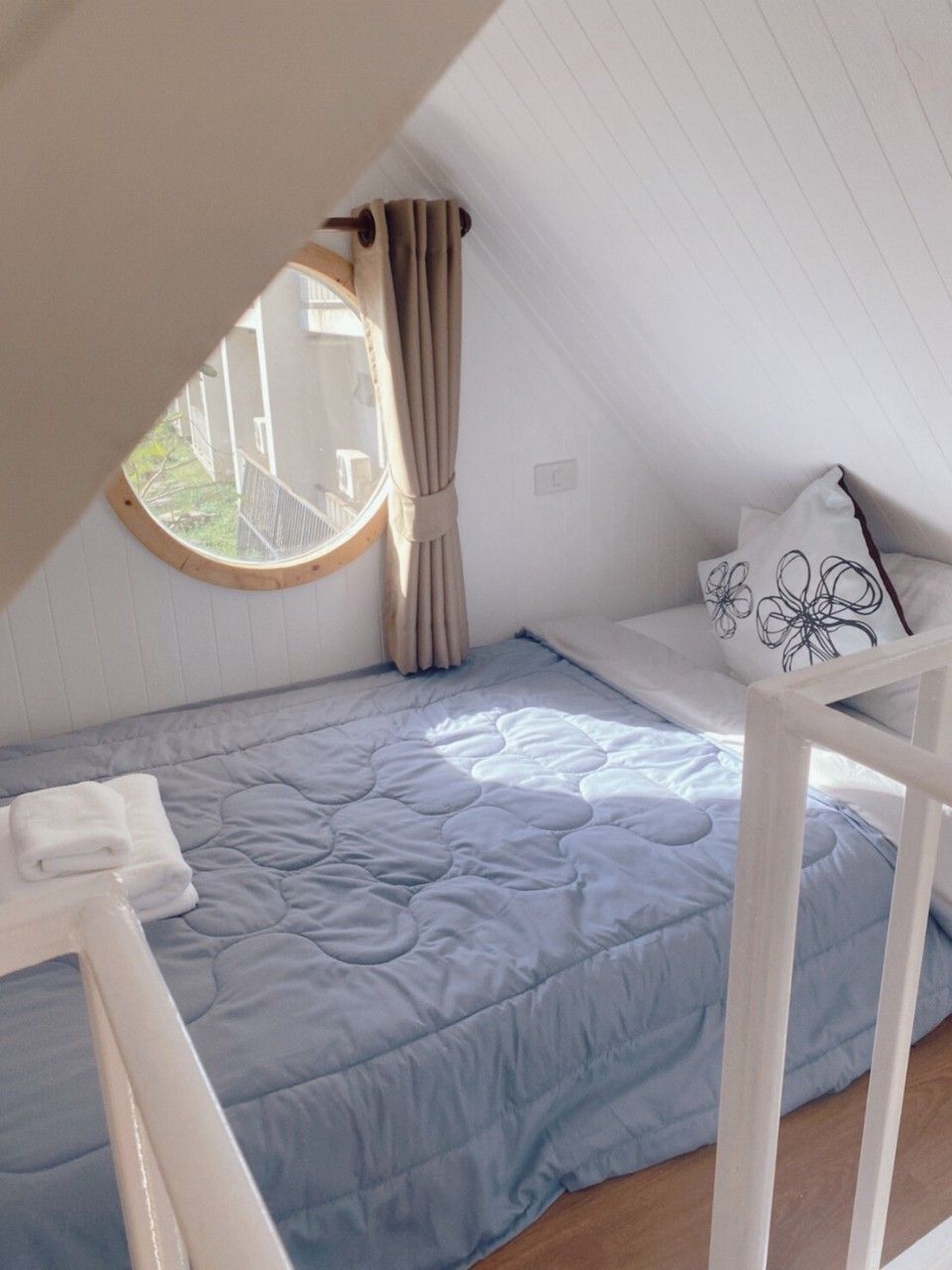 A single bed with a blue quilt in a room with a sloped ceiling and a window with curtains.