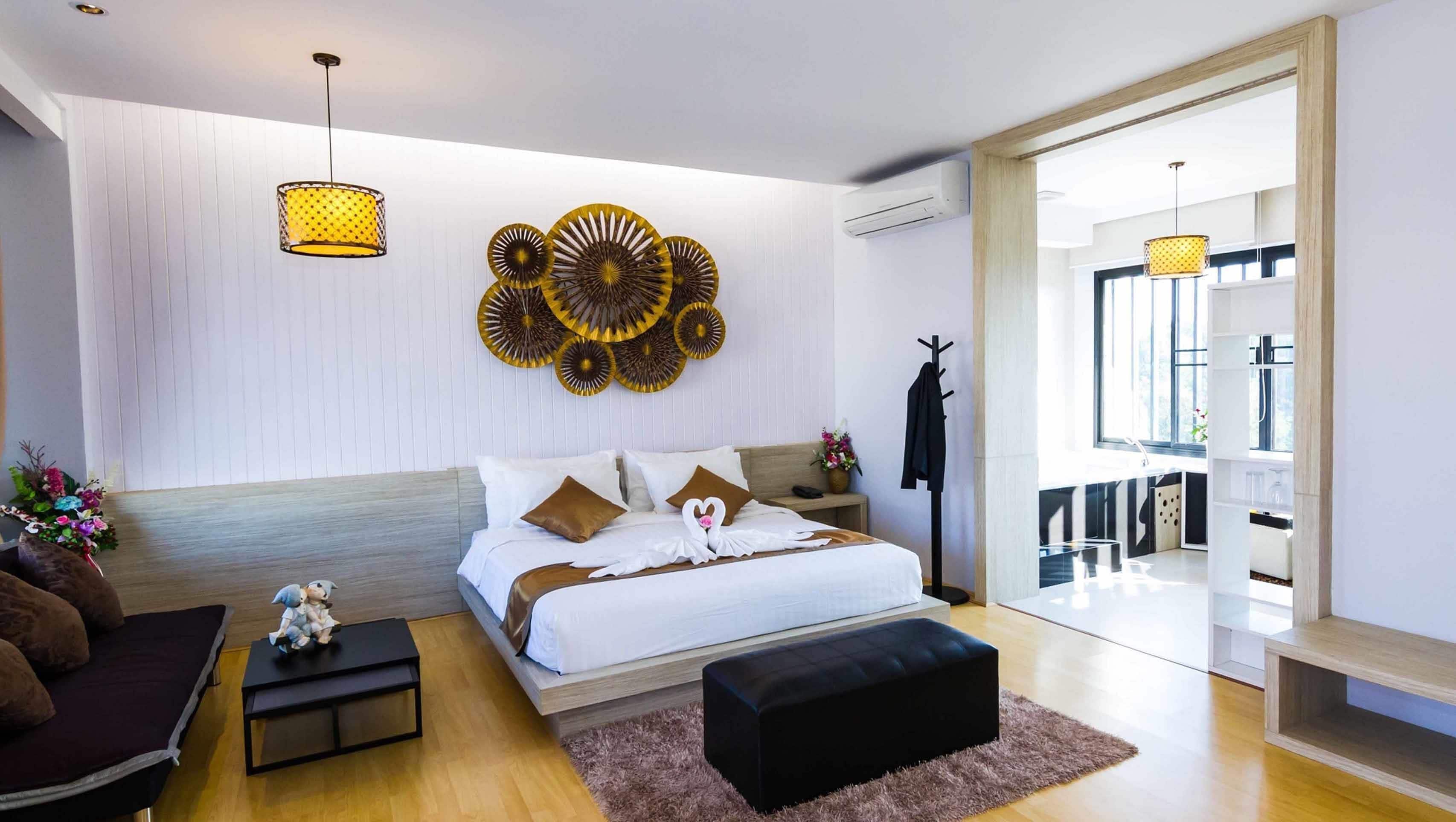 A modern bedroom with a large bed, a small side table, a black ottoman, a decorative wall piece, and a hanging light fixture.