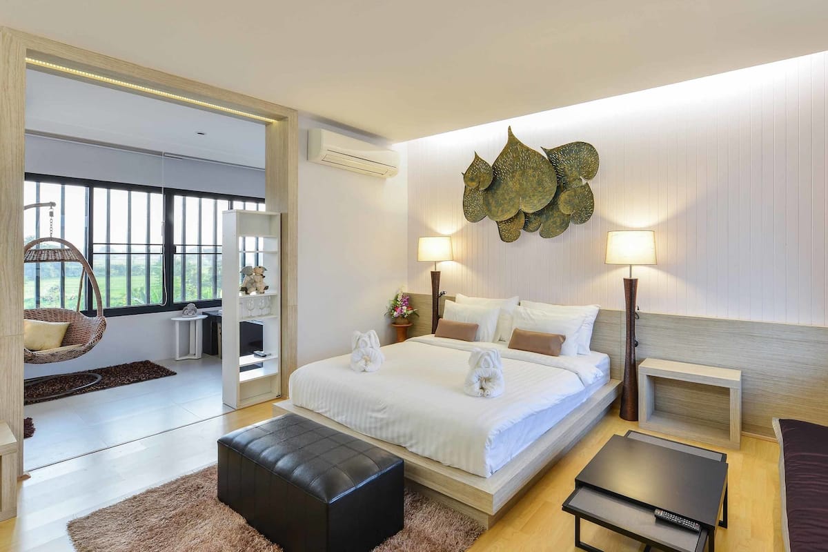 A modern hotel room with a large bed, bedside lamps, a small table, a sofa, and a view of greenery through the window.