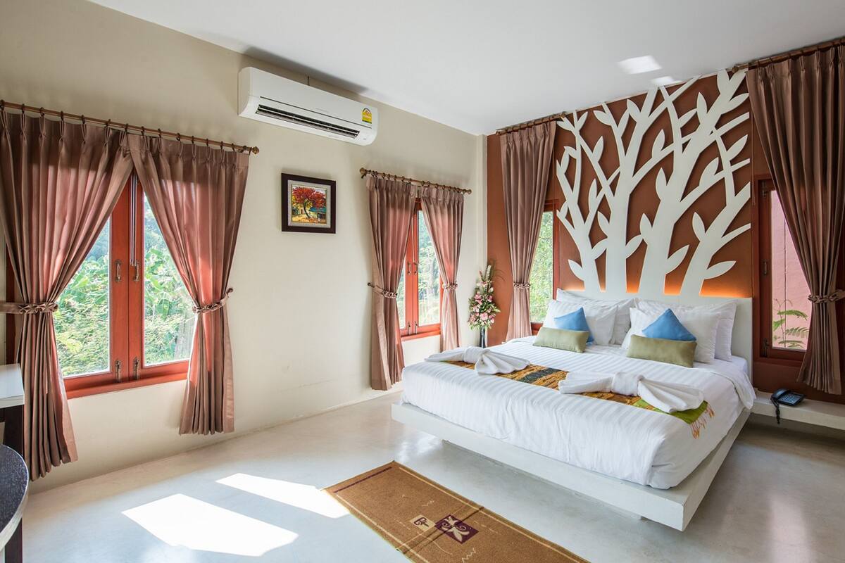 A bedroom with a large bed, two windows with curtains, an air conditioning unit, a wall-mounted decorative tree, and a small bedside table with a lamp.