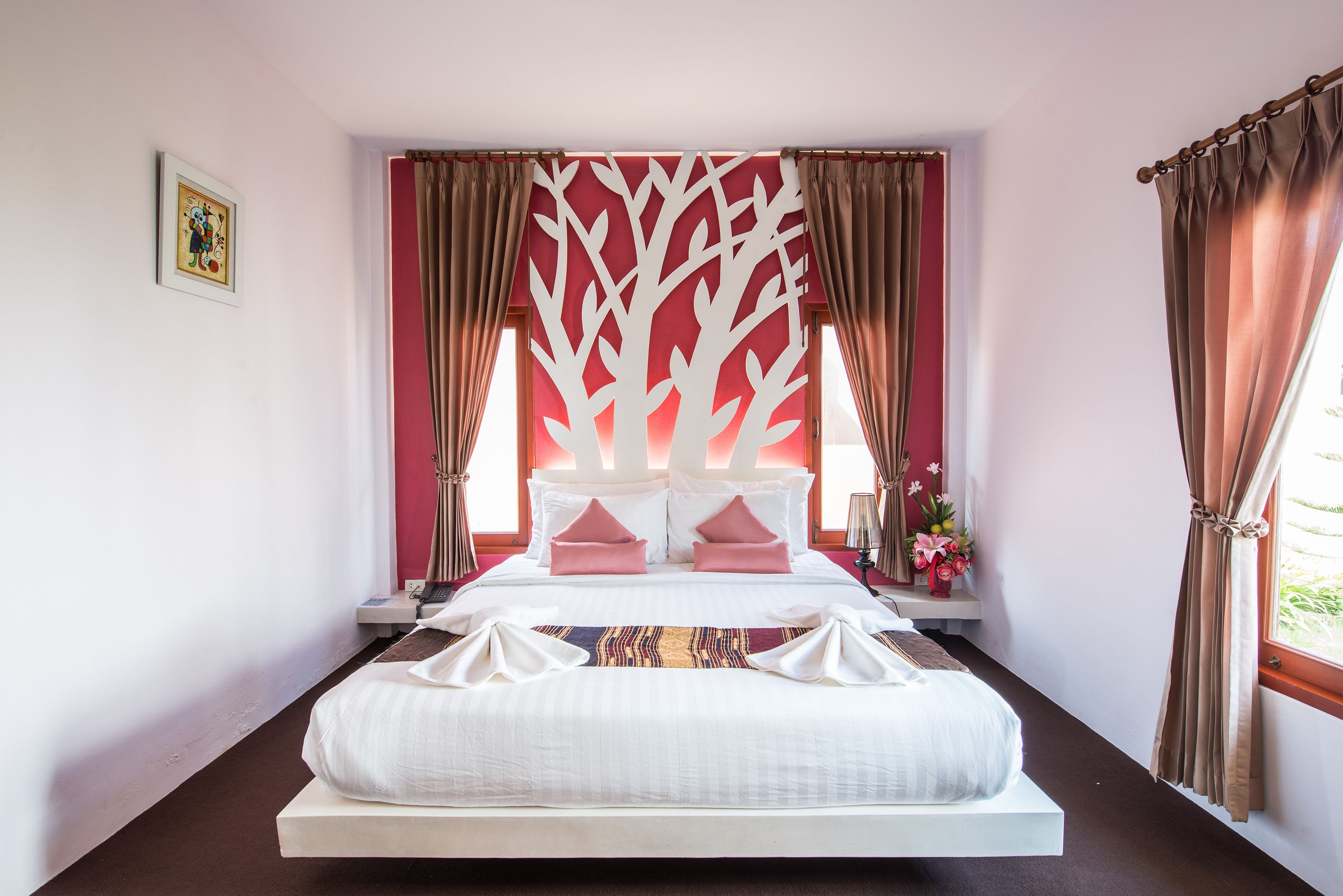 A neatly made bed with white linens and pink pillows, a red and white decorative wall panel, and a window with curtains.