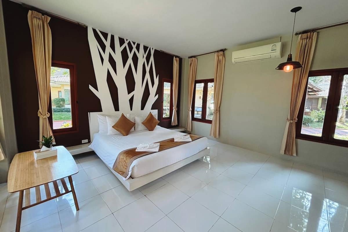 A bedroom with a large bed, a wooden nightstand, a wall-mounted air conditioner, and windows with curtains.