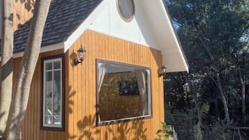 A small wooden house with a pitched roof, a large window, and potted plants around it.