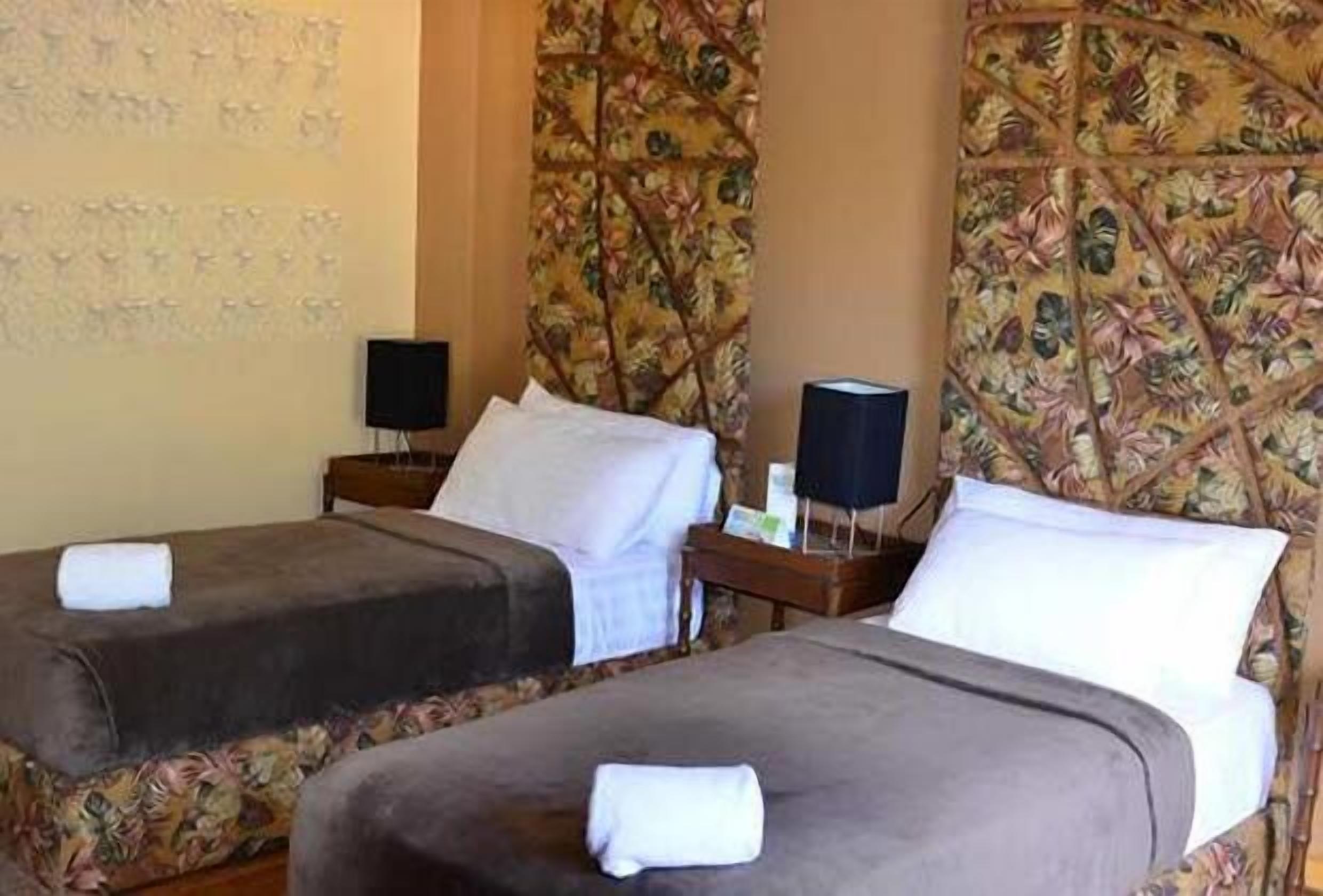 Camera d'albergo con due letti, ognuno con un cuscino e un asciugamano piegato. Due comodini con lampade e un tavolino con una bottiglia e un bicchiere.