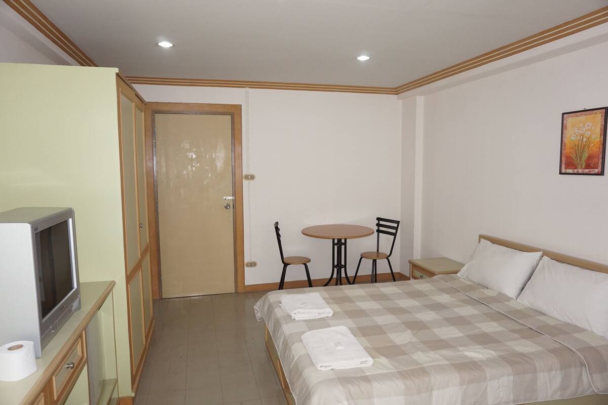 Habitación de hotel con cama, televisión, una mesita pequeña con dos sillas, una puerta y un cuadro en la pared.