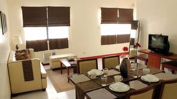 Un comedor con una mesa dispuesta para cuatro, un sofá, una televisión y ventanas amplias con persianas.