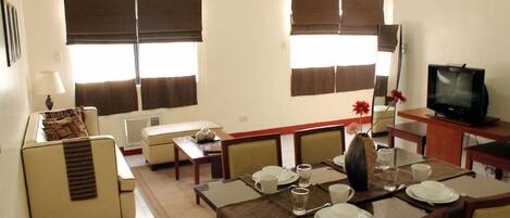 A dining area with a table set for four, a sofa, a television, and large windows with blinds.