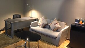 Una habitación de hotel moderna con un sofá gris, una mesita negra pequeña, un escritorio con una lámpara y un cuadro en la pared.