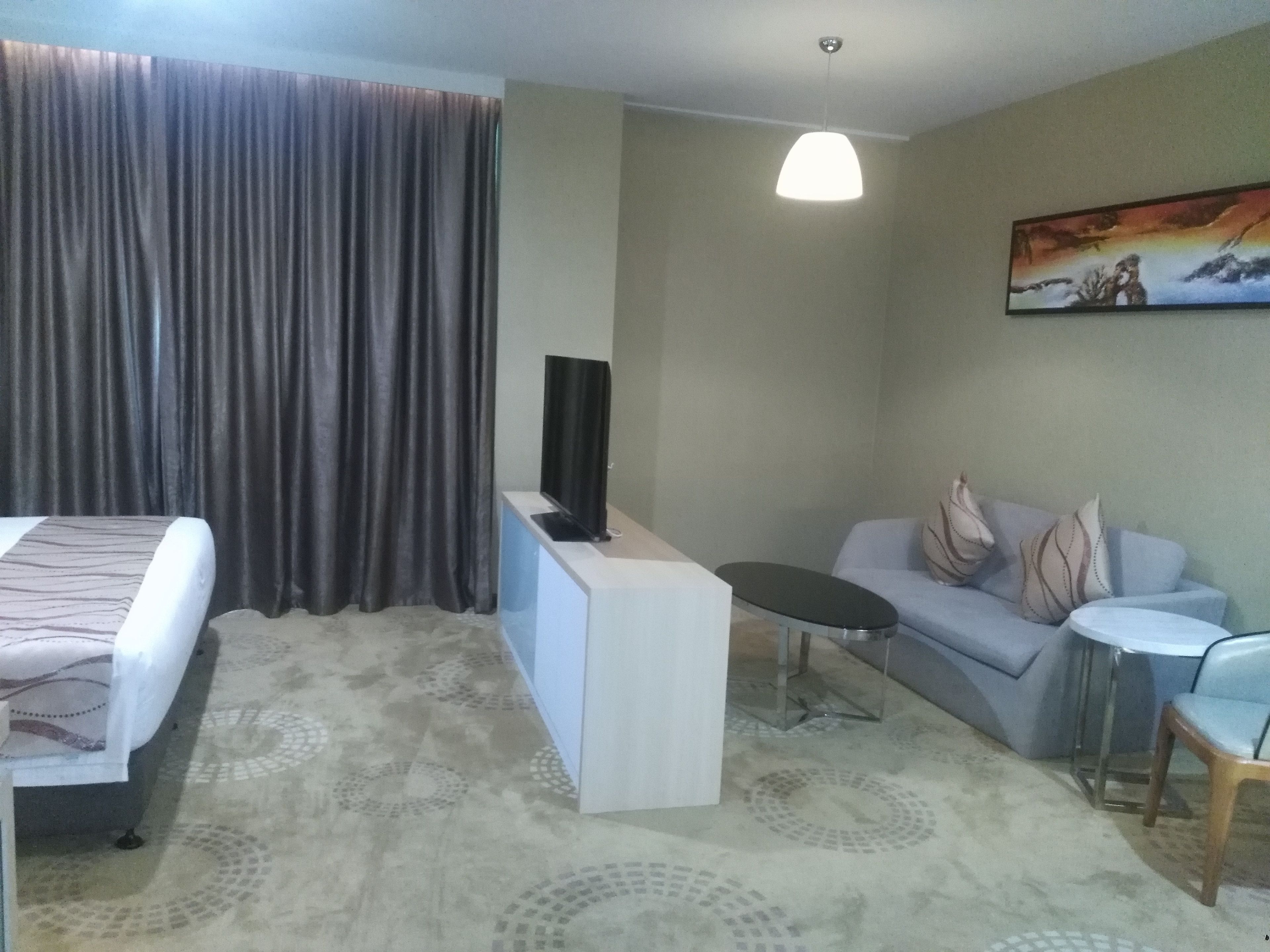 A hotel room with a bed, a TV on a stand, a small table, a sofa, and a painting on the wall.